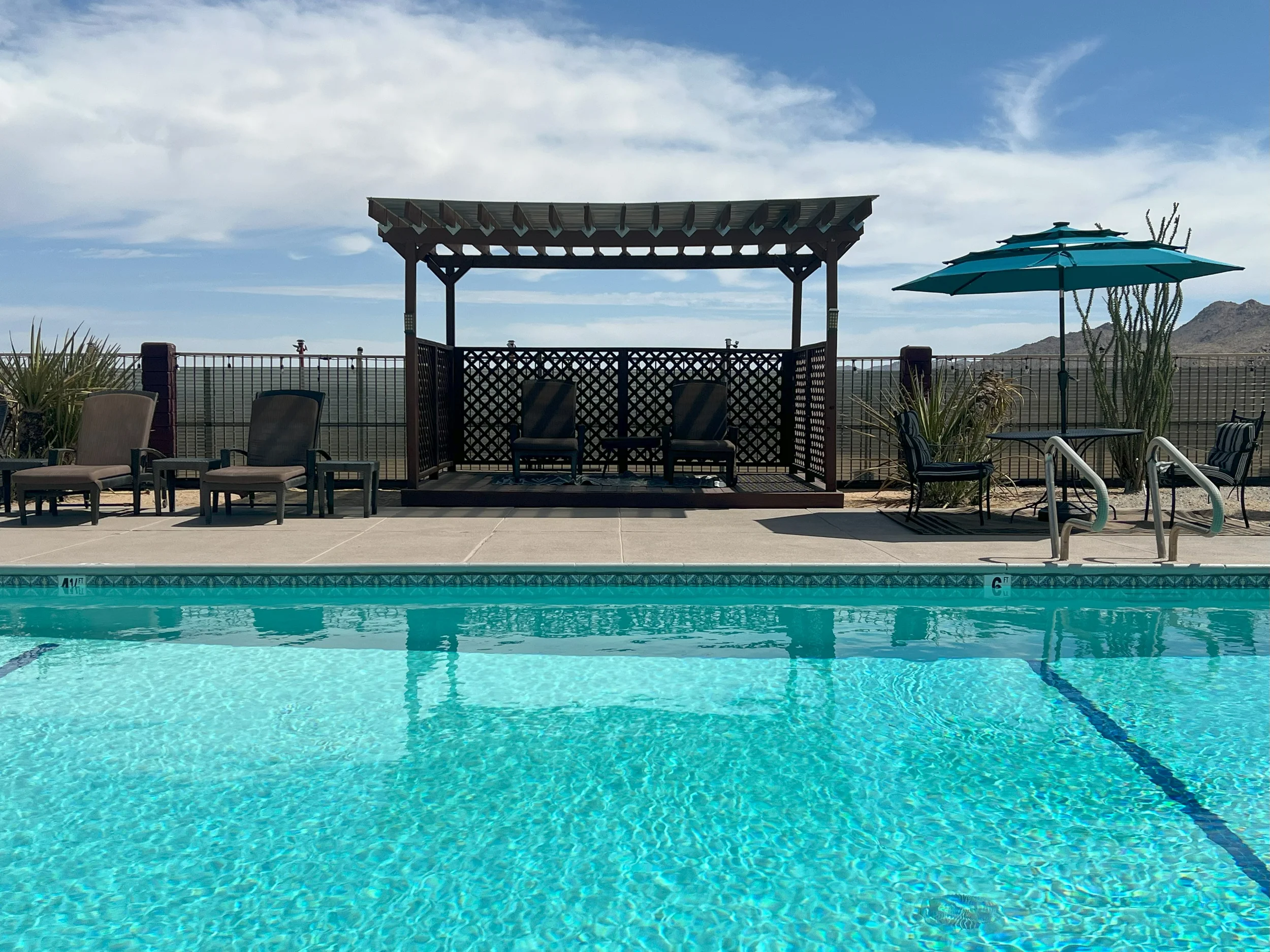 a pool with a wooden gazebo on the left and a table with an umbrella to the right
